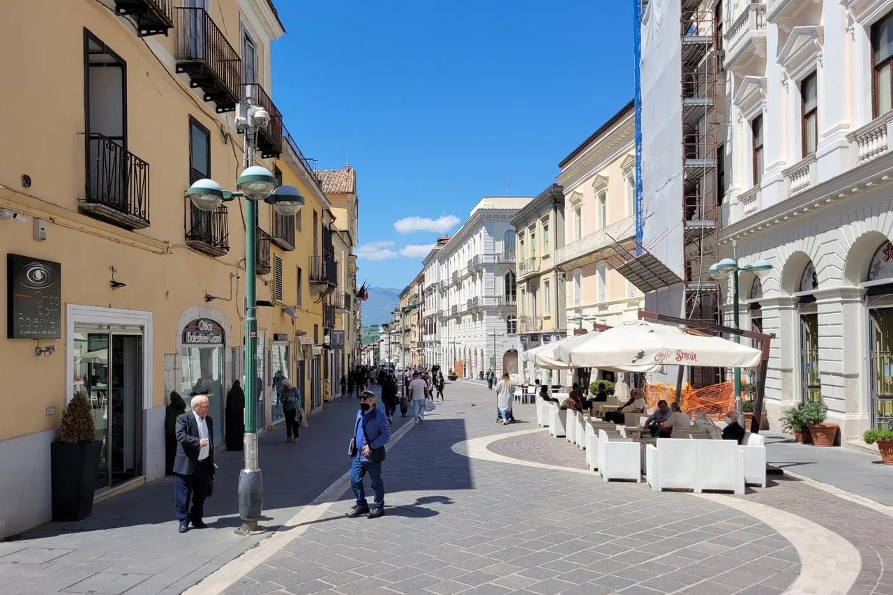 A sunny pedestrian street lined with shops and cafe tables